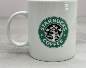 Può includere: Una tazza da caffè in ceramica bianca con il logo circolare verde e nero di Starbucks Coffee sulla parte anteriore. La tazza presenta una finitura liscia e lucida e un manico curvo sul lato sinistro, con l'iconica grafica della sirena.