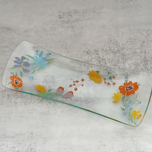May include: A clear glass serving tray with a rectangular shape and curved edges. The tray is decorated with colorful floral designs in orange, yellow, blue, and purple. The tray is approximately 12 inches long.