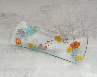 Puede incluir: Una bandeja de servir de vidrio transparente con forma rectangular y bordes curvos. La bandeja está decorada con diseños florales coloridos en naranja, amarillo, azul y morado. La bandeja mide aproximadamente 30 cm de largo.