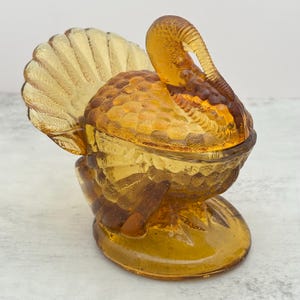 May include: An amber-colored glass turkey-shaped dish with a removable lid. The piece features detailed molded feather patterns on the body and a large, fanned-out tail. The turkey stands on a circular base, with the head curved downward.