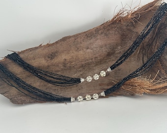 Può includere: Una collana di perline nere con accenti argentati e abbellimenti in strass trasparenti. La collana è a strati e ha più fili.