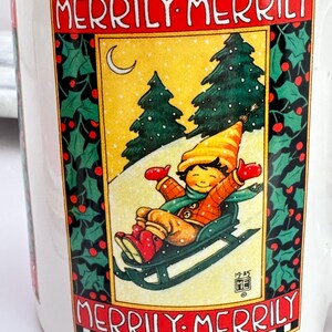 Puede incluir: Taza de cer&aacute;mica blanca con una escena invernal festiva. La taza presenta a un ni&ntilde;o desliz&aacute;ndose en trineo por una colina nevada, con las palabras "Merrily Merrily" en rojo. Acebo y bayas enmarcan la imagen, con un borde amarillo y negro.