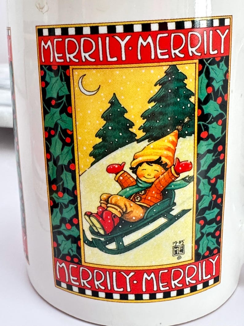 Puede incluir: Taza de cer&aacute;mica blanca con una escena invernal festiva. La taza presenta a un ni&ntilde;o desliz&aacute;ndose en trineo por una colina nevada, con las palabras "Merrily Merrily" en rojo. Acebo y bayas enmarcan la imagen, con un borde amarillo y negro.