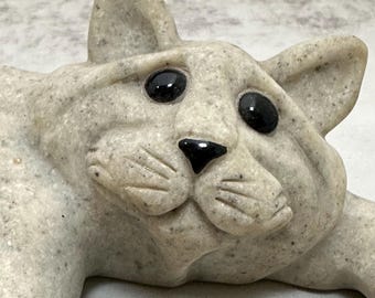 Può includere: Una statuetta di gatto in pietra color beige chiaro maculato, con due occhi neri lisci e lucidi e un piccolo naso nero triangolare. Il gatto presenta baffi scolpiti, orecchie a punta e una superficie ruvida che ricorda la pietra naturale.