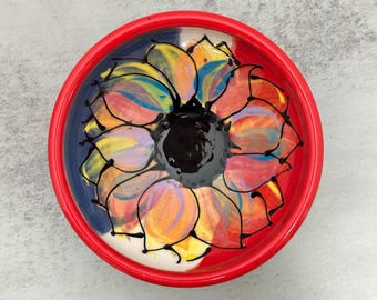 Può includere: Una ciotola rotonda in ceramica con un bordo rosso brillante. L'interno presenta un disegno floreale dipinto a mano con un centro nero solido circondato da petali sovrapposti in tonalità di giallo, arancione, rosa e blu, delineati in nero.