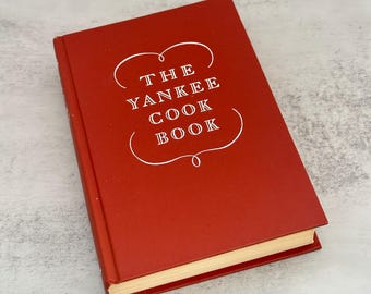 Puede incluir: Un libro de cocina rojo titulado "The Yankee Cook Book" en letras blancas. El libro está cerrado y apoyado sobre una superficie gris claro. El título está encerrado en un contorno blanco decorativo.