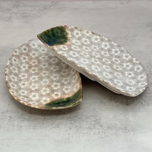 May include: Two oval ceramic dishes featuring a repeating white flower pattern on a light gray background. Each dish has a distinct green and blue glazed accent on one end, providing a contrast to the floral design and the neutral base.