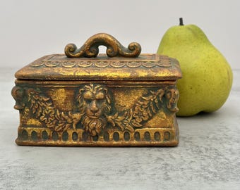 Puede incluir: Una caja rectangular de tono dorado con una tapa que presenta un relieve de cabeza de león, guirnaldas florales y un asa curva. La caja tiene un acabado desgastado con acentos oscuros y está colocada junto a una pera verde.
