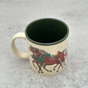 May include: A cream-colored ceramic mug with a dark forest green interior and handle. The exterior features a detailed illustration of two brown horses wearing festive green wreaths and red ribbons, set against a background of small dots.