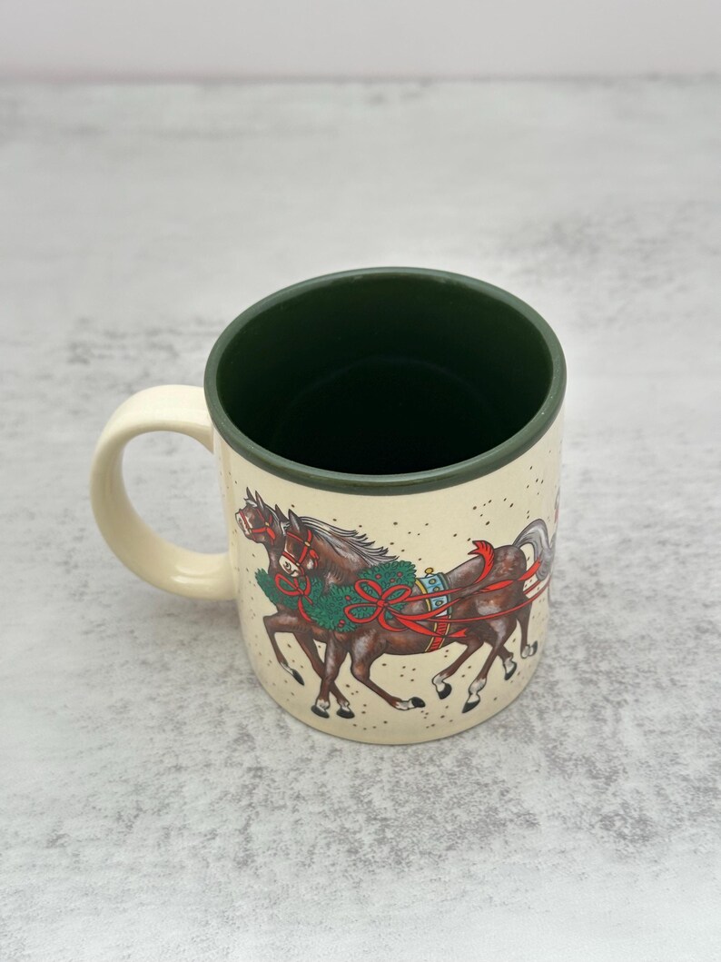 May include: A cream-colored ceramic mug with a dark forest green interior and handle. The exterior features a detailed illustration of two brown horses wearing festive green wreaths and red ribbons, set against a background of small dots.