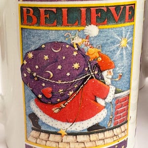 Puede incluir: Taza de cer&aacute;mica blanca con una ilustraci&oacute;n festiva. El dise&ntilde;o presenta a Pap&aacute; Noel en un tejado, llevando un saco de regalos, con la palabra &laquo;BELIEVE&raquo; encima. El borde tiene estrellas y lunas crecientes.