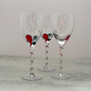 May include: Three clear glass wine glasses with long stems featuring a twisted design of red and green glass accents. The base of each bowl has a small red glass drop detail. The glasses have a circular foot and a smooth, transparent finish.