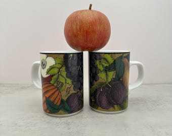 Può includere: Due tazze in ceramica bianca con una stampa grafica dettagliata di vari frutti, tra cui uva, prugne e mele in tonalità di verde, viola e arancione. Una singola mela rossa e gialla è bilanciata sopra le due tazze.