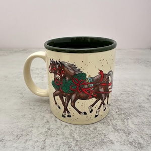 May include: A cream-colored ceramic mug with a dark green interior and rim. The exterior features a detailed illustration of two brown horses wearing green wreaths with red ribbons and bows, set against a background of small black speckles.