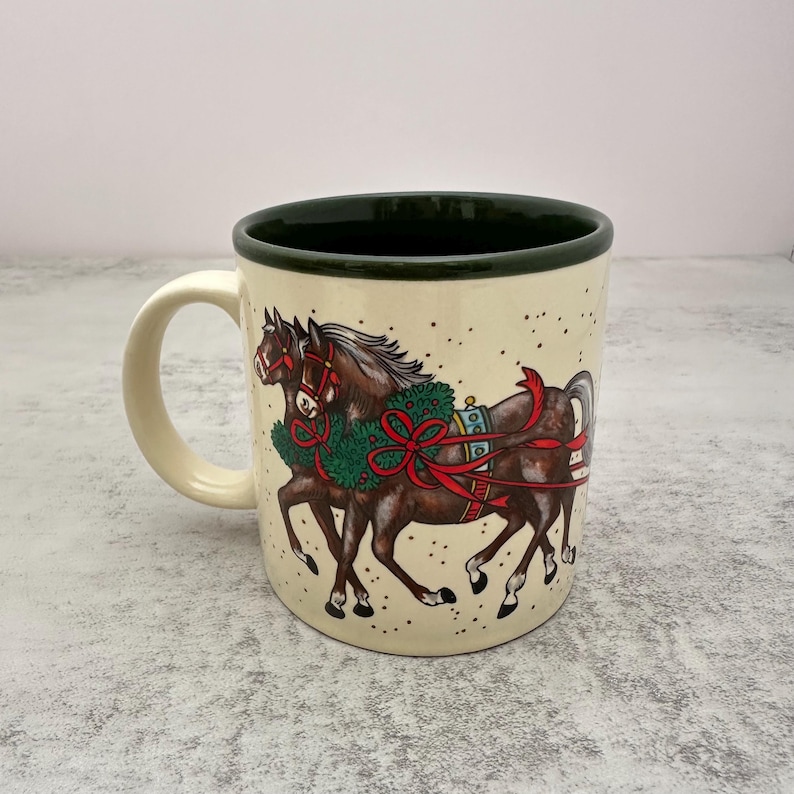 May include: A cream-colored ceramic mug with a dark green interior and rim. The exterior features a detailed illustration of two brown horses wearing green wreaths with red ribbons and bows, set against a background of small black speckles.
