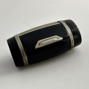 May include: A black, barrel-shaped container with silver-colored metal trim and a silver handle. The container has a glossy finish and appears to be made of a hard material. The design is simple and elegant.