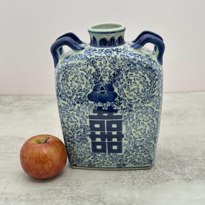 May include: A rectangular ceramic vase with a light blue and white finish, featuring dark blue floral scrollwork and a central double happiness character. The vase has two dark blue handles at the top and is shown next to a red apple for scale.