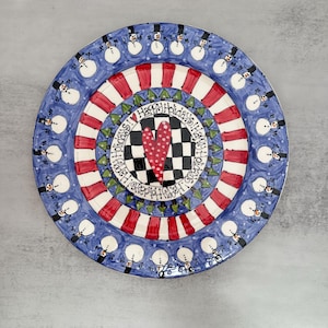 May include: A round ceramic plate featuring a blue border with a repeating pattern of snowmen wearing black top hats. The center displays a black and white checkered square with a red heart, surrounded by red and white stripes and text reading Happy Holidays.