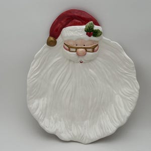 May include: A ceramic Santa Claus-shaped serving platter. The platter features a red hat with a gold bell, a white beard, and a smiling face with glasses. The platter is white with a glossy finish.