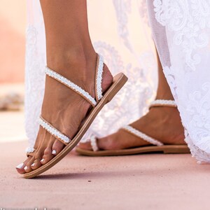 May include: White beaded sandals with a brown leather strap and sole. The sandals are worn by a person wearing a white lace dress.