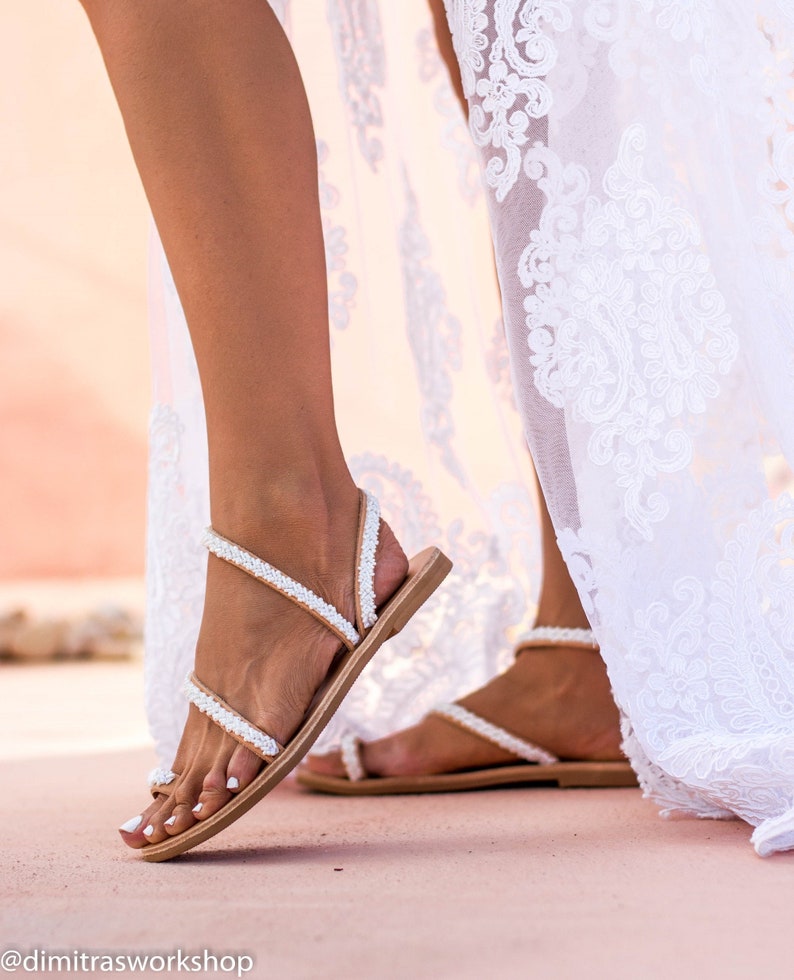 May include: White beaded sandals with a brown leather strap and sole. The sandals are worn by a person wearing a white lace dress.