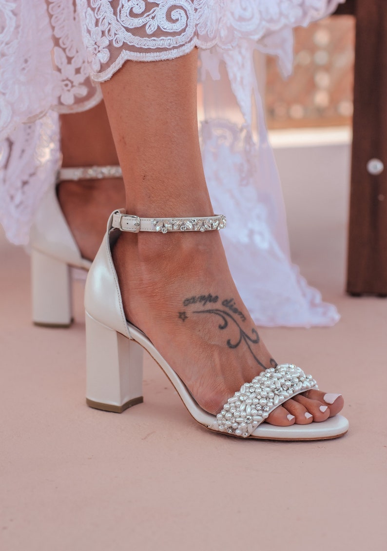 Puede incluir: Tacones de novia blancos con un tac&oacute;n grueso y un adorno de perlas y pedrer&iacute;a. Los tacones tienen una correa con pedrer&iacute;a alrededor del tobillo.