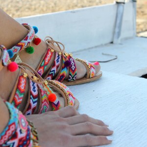 May include: Colorful, handmade sandals with intricate woven straps in vibrant patterns. The sandals feature pom-poms, gold chain accents, and leather straps. The design incorporates a variety of colors, including red, blue, green, and yellow.