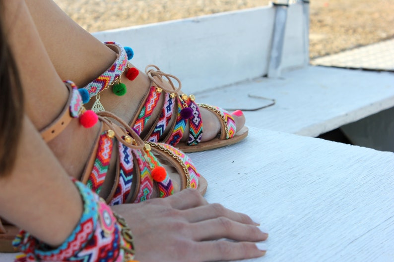 May include: Colorful, handmade sandals with intricate woven straps in vibrant patterns. The sandals feature pom-poms, gold chain accents, and leather straps. The design incorporates a variety of colors, including red, blue, green, and yellow.