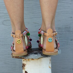 May include: A pair of leather sandals with colorful woven straps and pom-pom accents. Each sandal features a peace sign charm and a decorative chain with leaf pendants. The sandals have adjustable ankle straps.