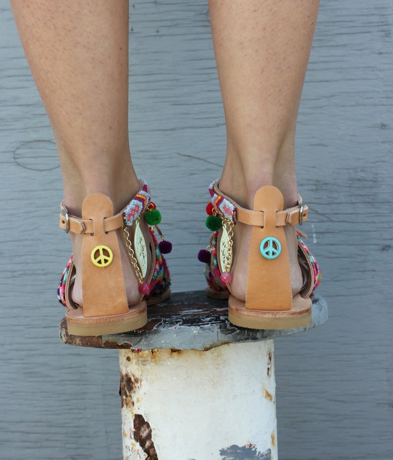 May include: A pair of leather sandals with colorful woven straps and pom-pom accents. Each sandal features a peace sign charm and a decorative chain with leaf pendants. The sandals have adjustable ankle straps.