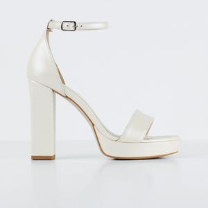Puede incluir: Sandalia de tacón alto con plataforma de color marfil, con tacón de bloque grueso y correa en el tobillo. La sandalia presenta una sola correa sobre los dedos y un cierre de hebilla. El tacón mide unos 12,7 cm.