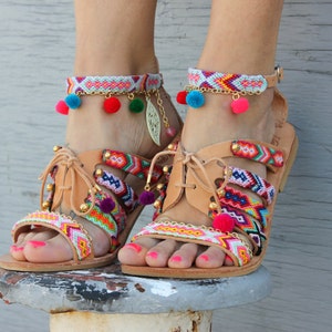 May include: Tan leather sandals with colorful woven straps and ankle bracelets. The sandals feature a multi-colored pattern with pom-poms and gold accents. The ankle bracelets have a similar design with pom-poms and gold chain details.