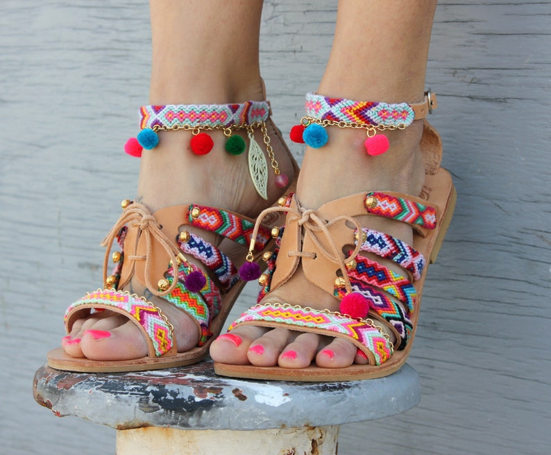 May include: Tan leather sandals with colorful woven straps and ankle bracelets. The sandals feature a multi-colored pattern with pom-poms and gold accents. The ankle bracelets have a similar design with pom-poms and gold chain details.