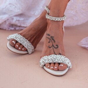 Puede incluir: Un par de sandalias de novia blancas con adornos de perlas. Las sandalias tienen una delicada correa de tobillo con pedrer&iacute;a.