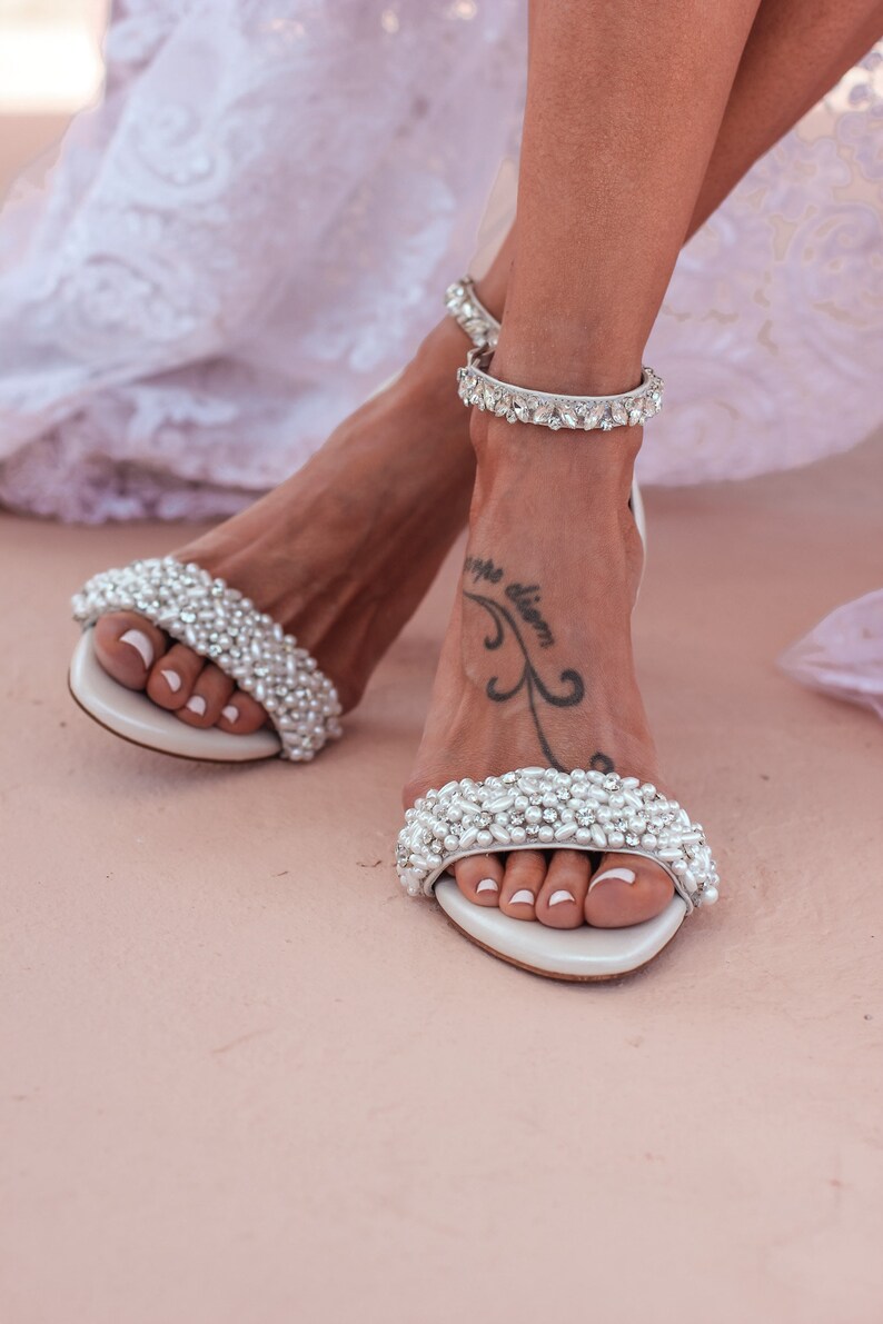 Puede incluir: Un par de sandalias de novia blancas con adornos de perlas. Las sandalias tienen una delicada correa de tobillo con pedrer&iacute;a.