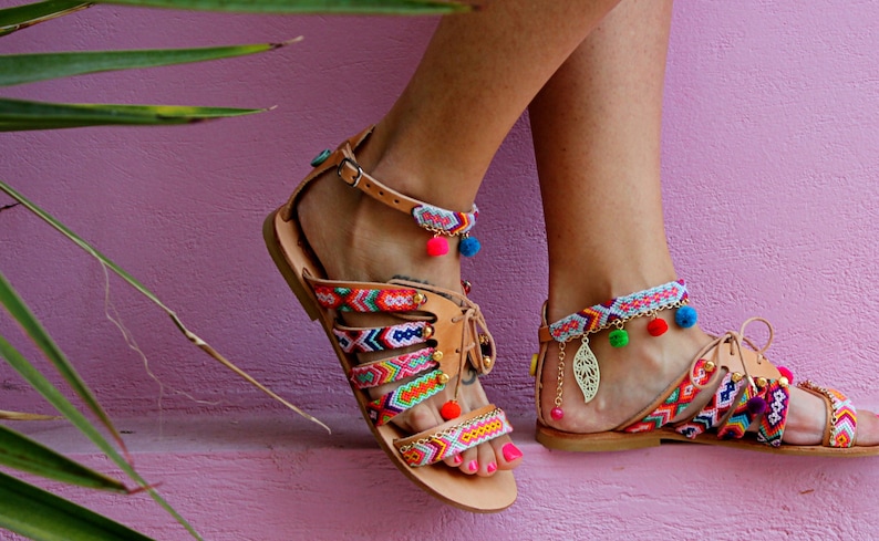 May include: Tan leather sandals with colorful woven straps and ankle straps. The sandals feature vibrant patterns in red, orange, green, and blue. Decorative pom-poms and gold charms add detail. The sandals are displayed against a pink background.