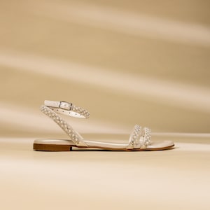K&ouml;nnte beinhalten: Wei&szlig;e Ledersandalen mit Perlenbesatz an den Riemen. Die Sandalen haben einen Schnallenverschluss und eine flache Sohle.