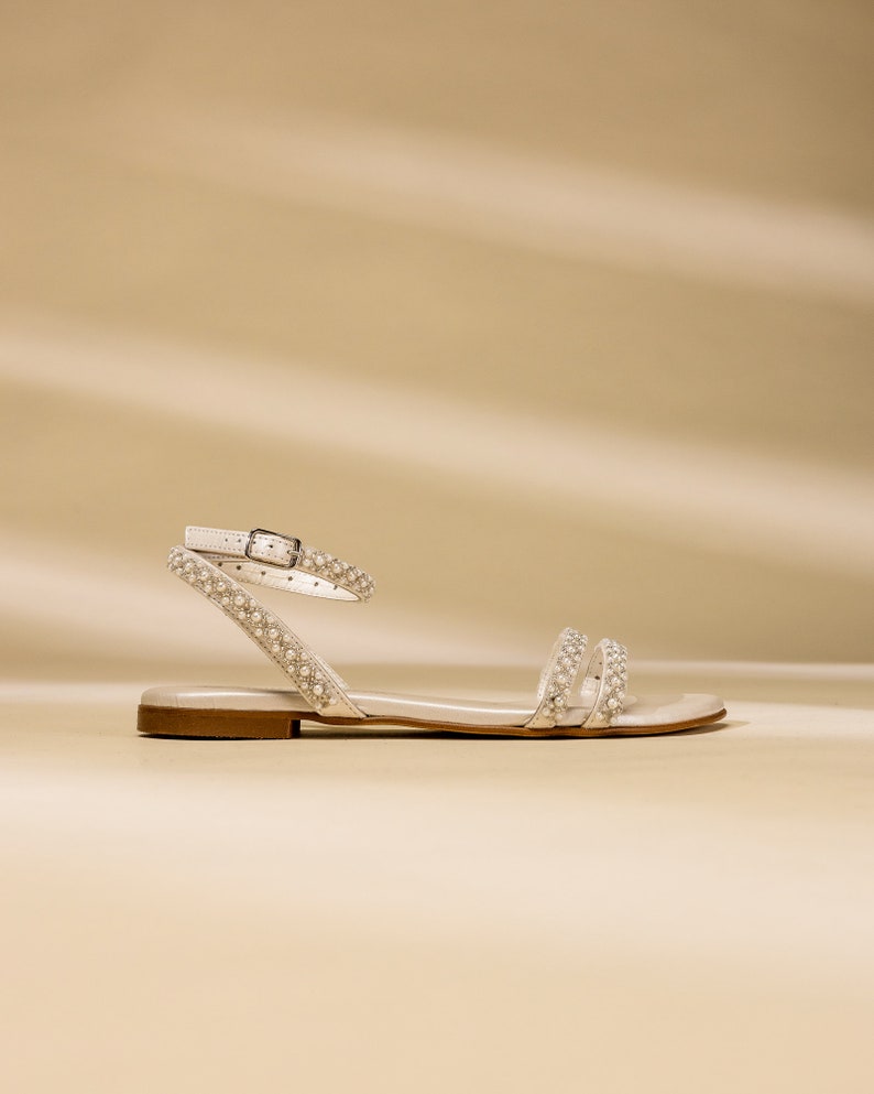 K&ouml;nnte beinhalten: Wei&szlig;e Ledersandalen mit Perlenbesatz an den Riemen. Die Sandalen haben einen Schnallenverschluss und eine flache Sohle.
