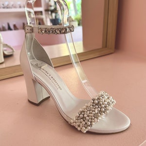 Puede incluir: Sandalias blancas de sat&eacute;n con tac&oacute;n y adorno de perlas y pedrer&iacute;a. Las sandalias tienen un tac&oacute;n grueso de bloque y una tira para el tobillo con una hebilla de pedrer&iacute;a. El nombre de la marca "Nitra's Workshop Handcrafted Sandals" est&aacute; impreso en la suela.