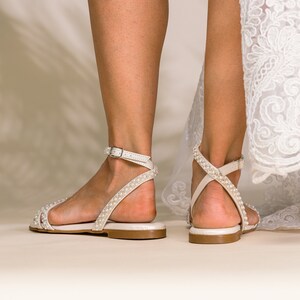 K&ouml;nnte beinhalten: Ein Paar elfenbeinfarbene Braut-Sandalen mit Perlenbesatz und Kn&ouml;chelriemen.
