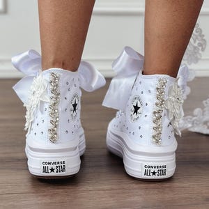 Peut inclure: Baskets montantes Converse All Star blanches avec semelles compensées, ornées de strass, de motifs floraux et de grands nœuds en satin blanc. Les baskets arborent le logo Converse All Star sur le talon.