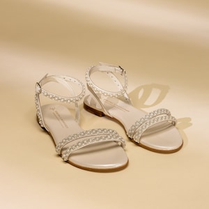K&ouml;nnte beinhalten: Ein Paar wei&szlig;e Ledersandalen mit Perlenbesatz. Die Sandalen haben einen d&uuml;nnen Riemen &uuml;ber den Zehen und einen Kn&ouml;chelriemen mit einer Schnalle. Die Sandalen sind von der Marke "Sophia Webster".