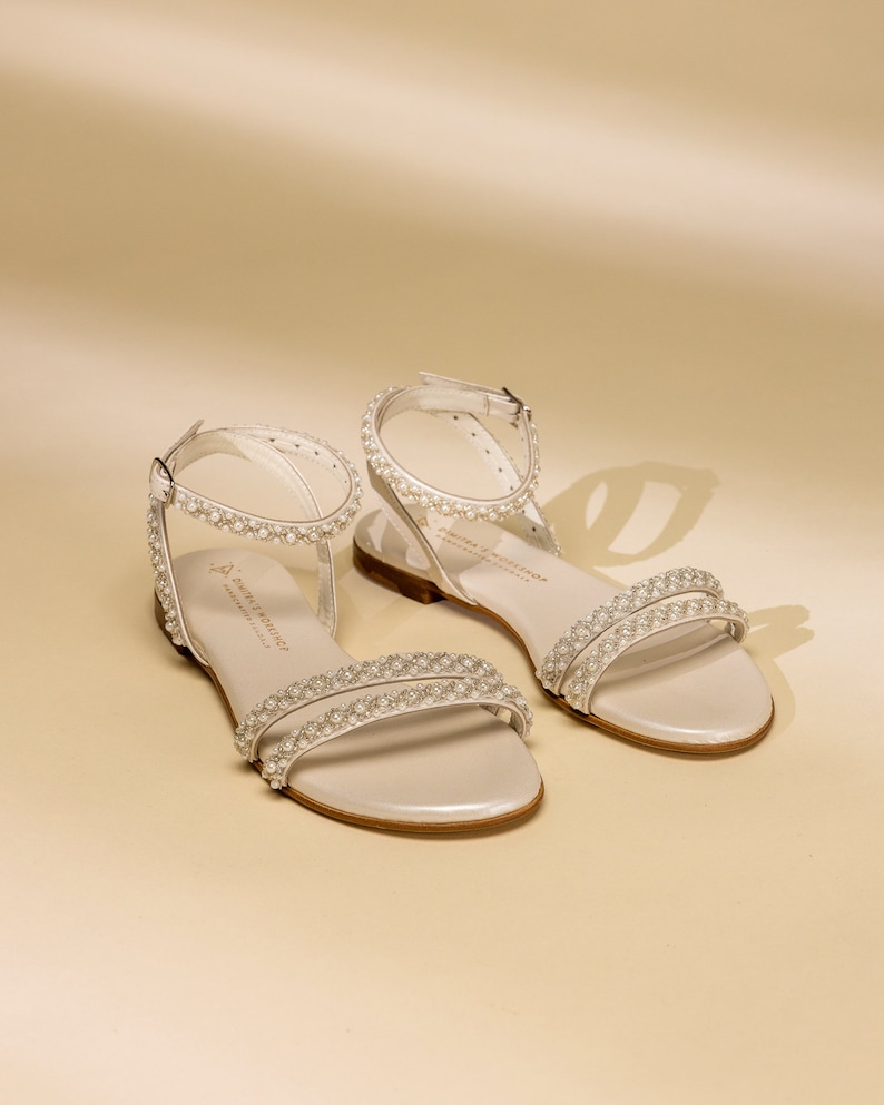 K&ouml;nnte beinhalten: Ein Paar wei&szlig;e Ledersandalen mit Perlenbesatz. Die Sandalen haben einen d&uuml;nnen Riemen &uuml;ber den Zehen und einen Kn&ouml;chelriemen mit einer Schnalle. Die Sandalen sind von der Marke "Sophia Webster".
