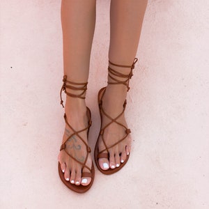 Op de afbeelding: Bruine leren sandalen met veters. De sandalen hebben een platte zool en zijn aan de enkel gebonden.