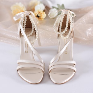 Puede incluir: Sandalias de tiras color marfil con detalles de perlas y lazos. Los tacones abiertos presentan múltiples tiras finas y un tacón de altura media. Delicadas hebras de perlas cuelgan de las correas del tobillo, añadiendo elegancia al calzado.