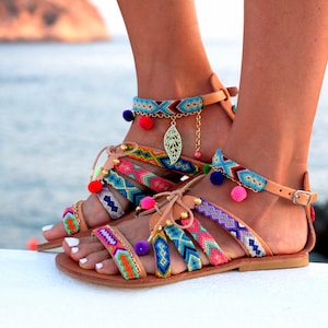 May include: Colorful gladiator sandals with woven straps in various colors, including pink, blue, and green. The sandals feature small pom-poms and a gold-colored leaf charm. The leather sandals have a buckle closure and are perfect for summer.