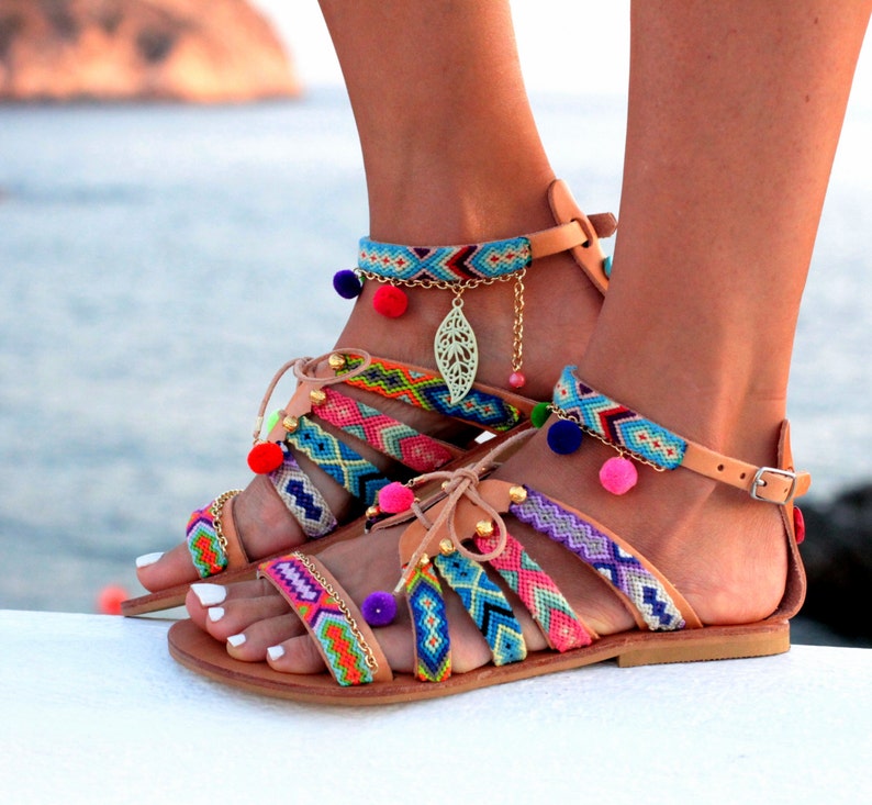 May include: Colorful gladiator sandals with woven straps in various colors, including pink, blue, and green. The sandals feature small pom-poms and a gold-colored leaf charm. The leather sandals have a buckle closure and are perfect for summer.