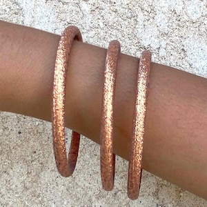 May include: Three copper-colored bangles with a textured surface are stacked on a wrist. The bangles have a metallic sheen and a simple, minimalist design. The jewelry is displayed against a neutral background.