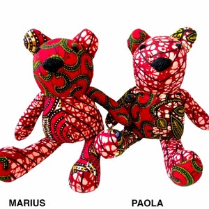 May include: Two stuffed teddy bears made from red fabric with intricate yellow, black, and white patterns. The bear on the left is labeled MARIUS, and the bear on the right is labeled PAOLA. Both bears feature black stitched noses and ears.