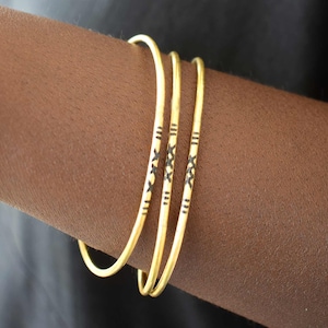 Op de afbeelding: Drie dunne goudkleurige metalen armbanden met een herhalend zwart geëtst patroon van kleine kruisjes en verticale lijnen. Deze ronde armbanden hebben een gladde, gepolijste afwerking en worden samen als een set op de onderarm gedragen.